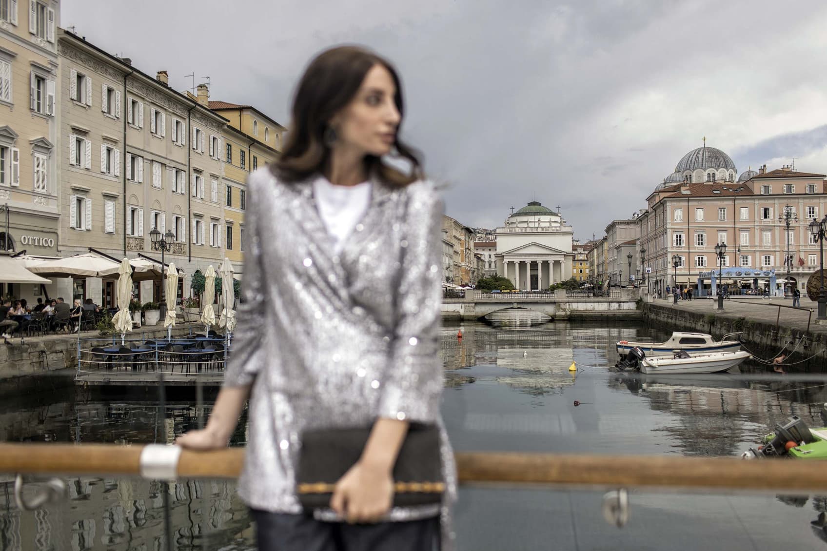 A woman at the harbour is watching towards the boats. Her body is blurred out but you can make out that she is dressed very fancy. In the back you can see the historic city of Trieste and its beautiful houses.