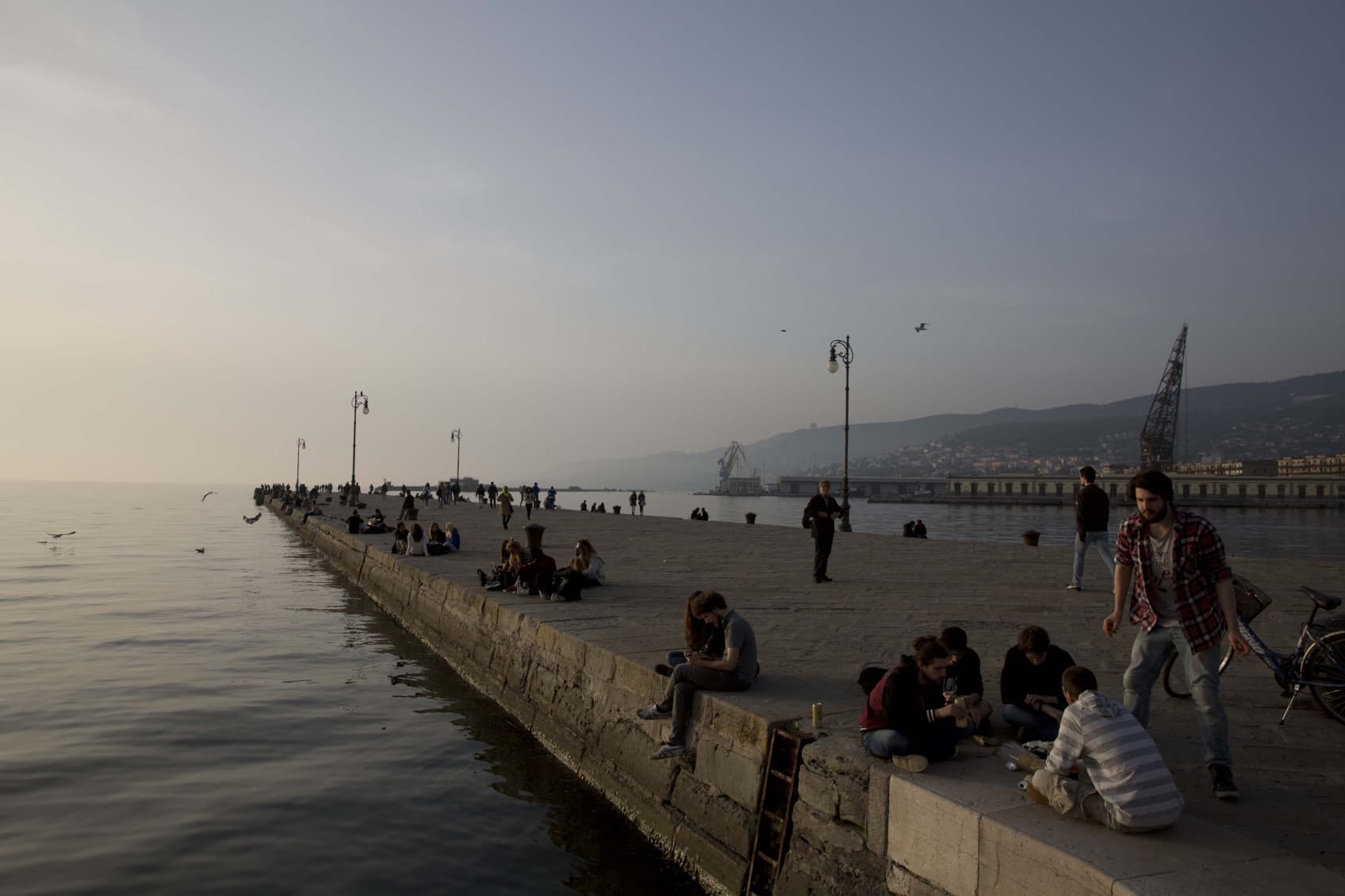 Auf dem Bild ist der Hafen von Triest zu sehen, mit Blick auf das Meer. Dort sitzen Menschen am Hafen und genießen den Sonnenuntergang.