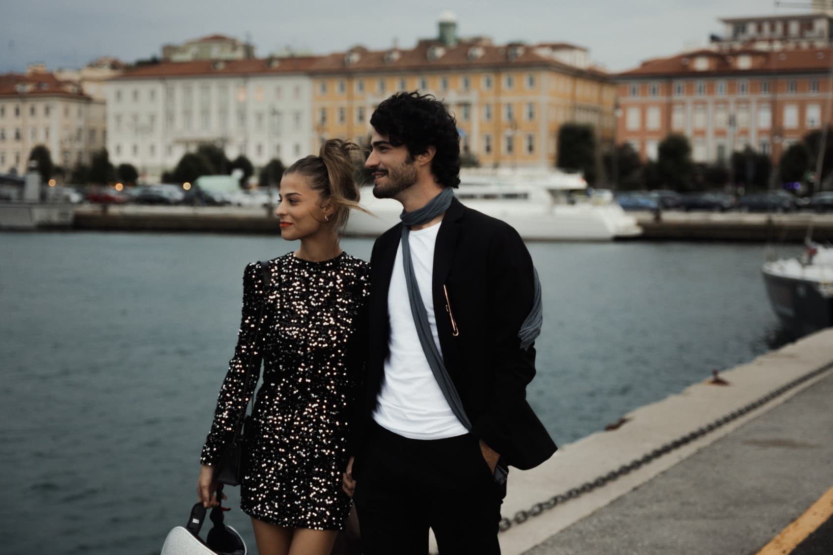 A young couple, dressed in their Sunday best, is strolling along the harbor in Trieste, walking away from URBANAUTS STUDIOS Minelli. To their left is the harbor wall, and in the background, you can see yachts anchored in the harbor.