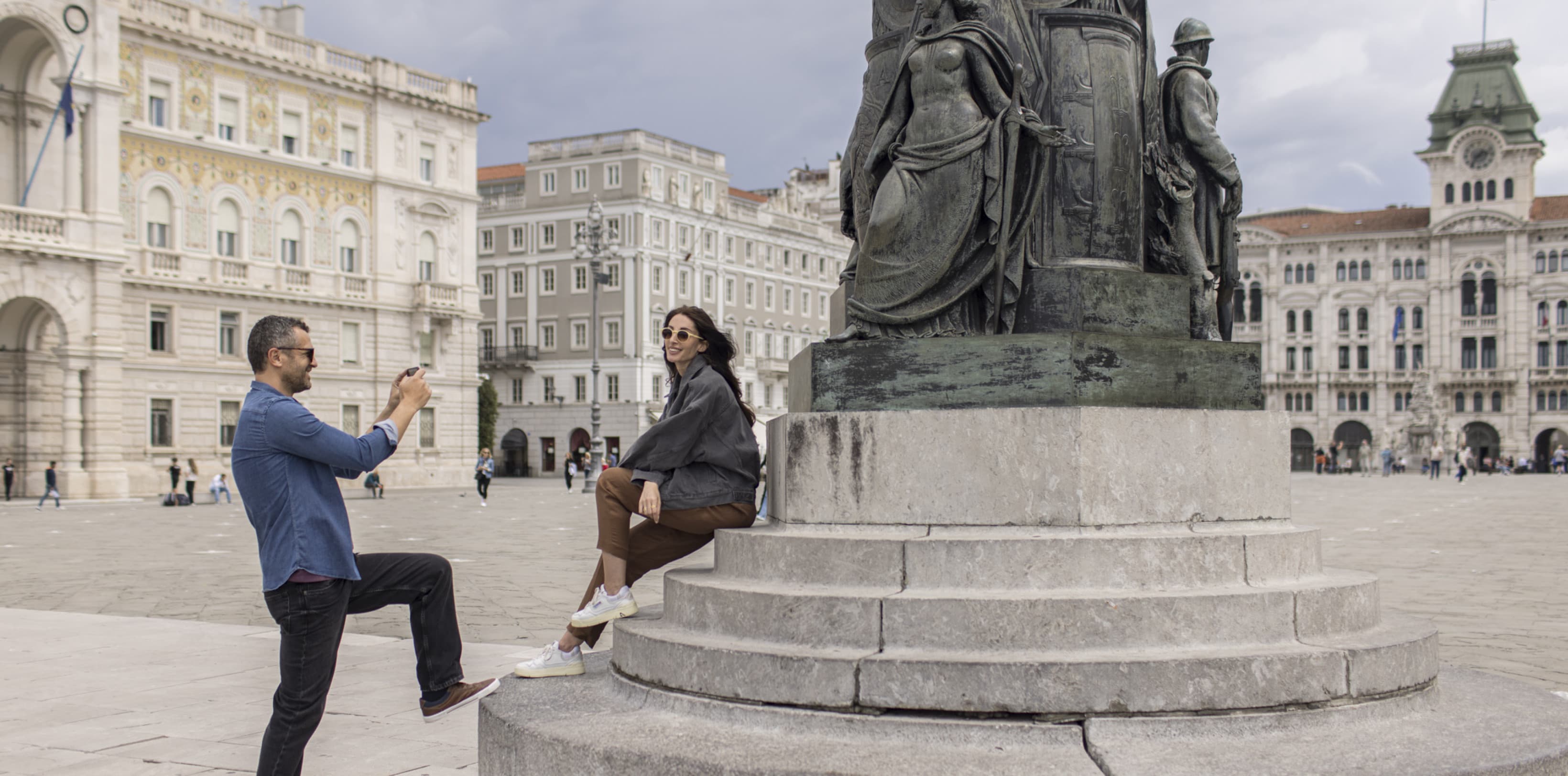 A man is taking a picture of a woman at the main Square in Trieste in front of a statue.