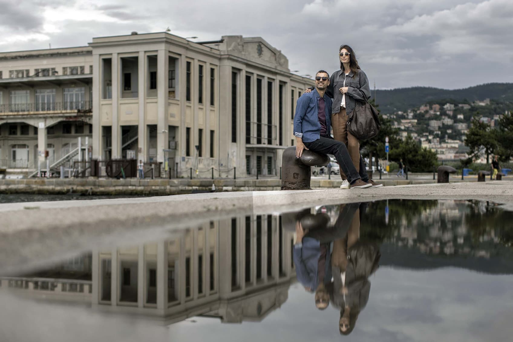 A couple is posing in a touristic picture in front of a warehouse at the Trieste Harbour. Their image is reflecting in a rain puddle.