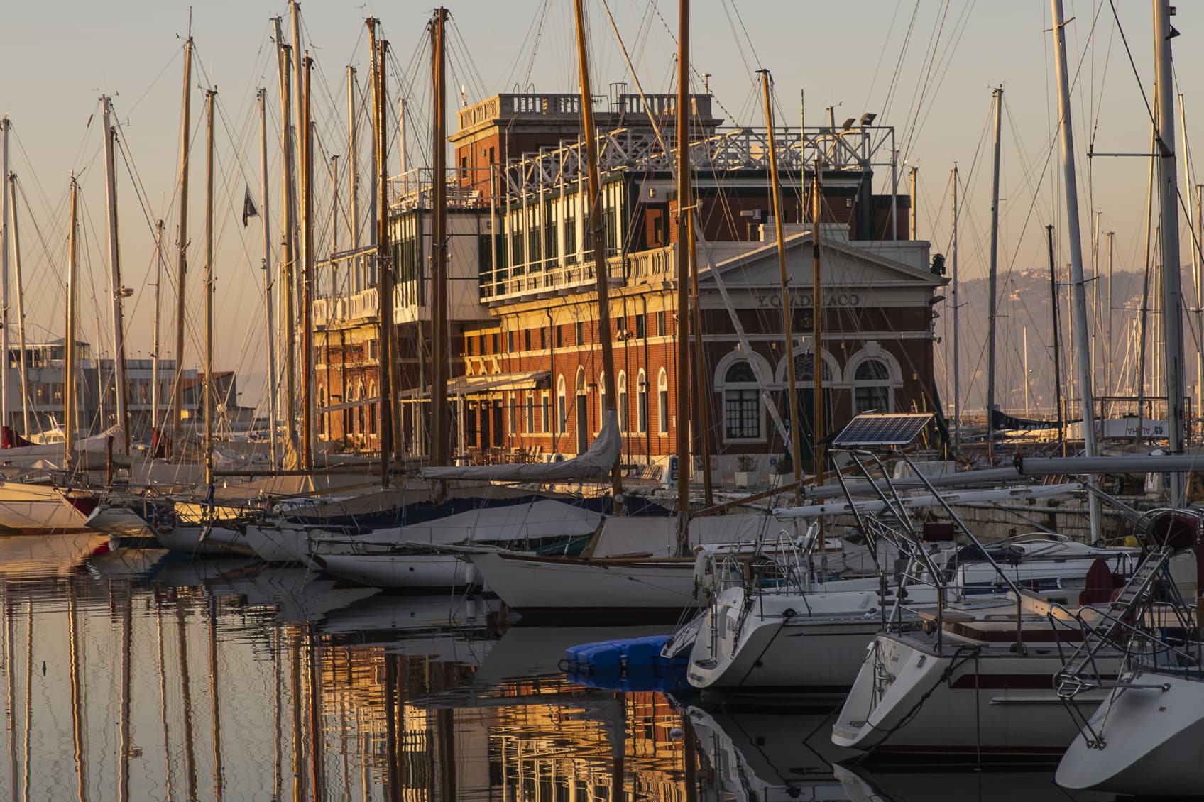 Blick auf die vielen Segelboote im Hafen von Triest bei Sonnenuntergang, mit einem Lagerhaus im Hintergrund.