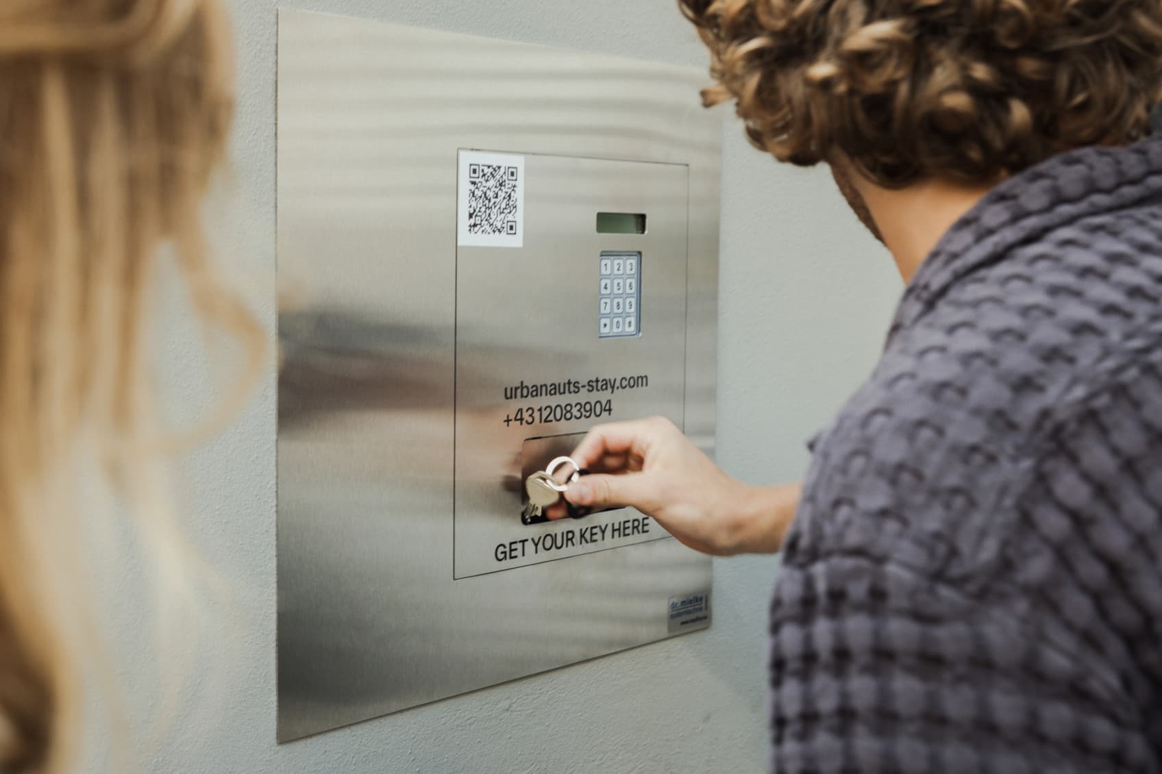 Two guests are checking in at any URBANAUTS location and recieved their Check In information beforehand. At 3 pm they can use the code to receive the key. The guy is grabbing the key from a little hatch below the keypad. All contact information is also shown on the safe.