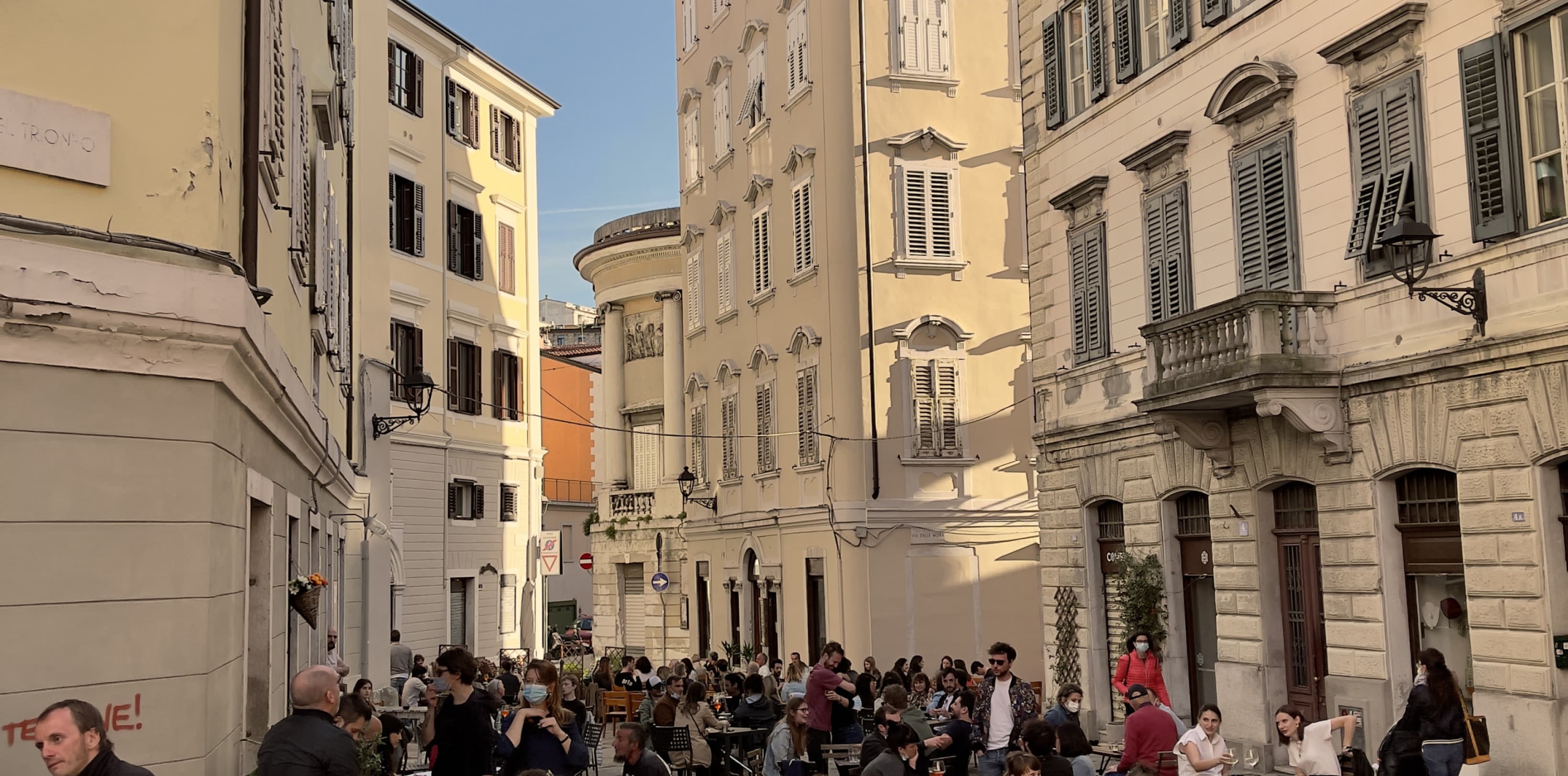 View into the busy streets of Trieste. The people are sitting outside on chairs enjoying a drink in the sun. In the background the historic buildings of Trieste are shown.