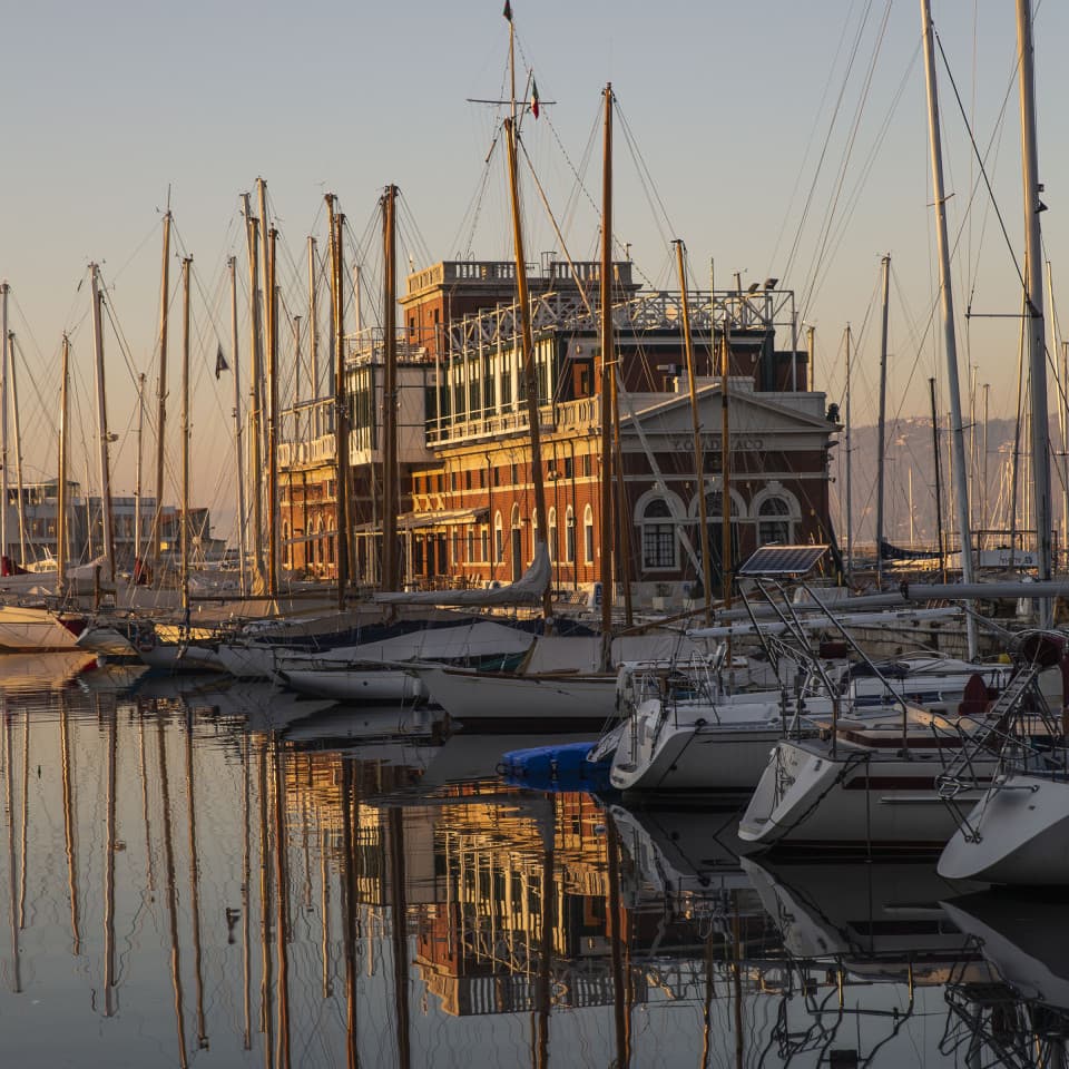 View of the many sailing boats in Trieste harbor at sunset, with a warehouse in the background.