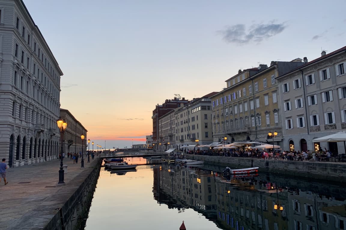 Das Bild zeigt einen Blick auf den Hafen von Triest am Abend. Auf der rechten Seite sind historische Häuser und Restaurants zu sehen, während die Sonne über dem Wasser untergeht.