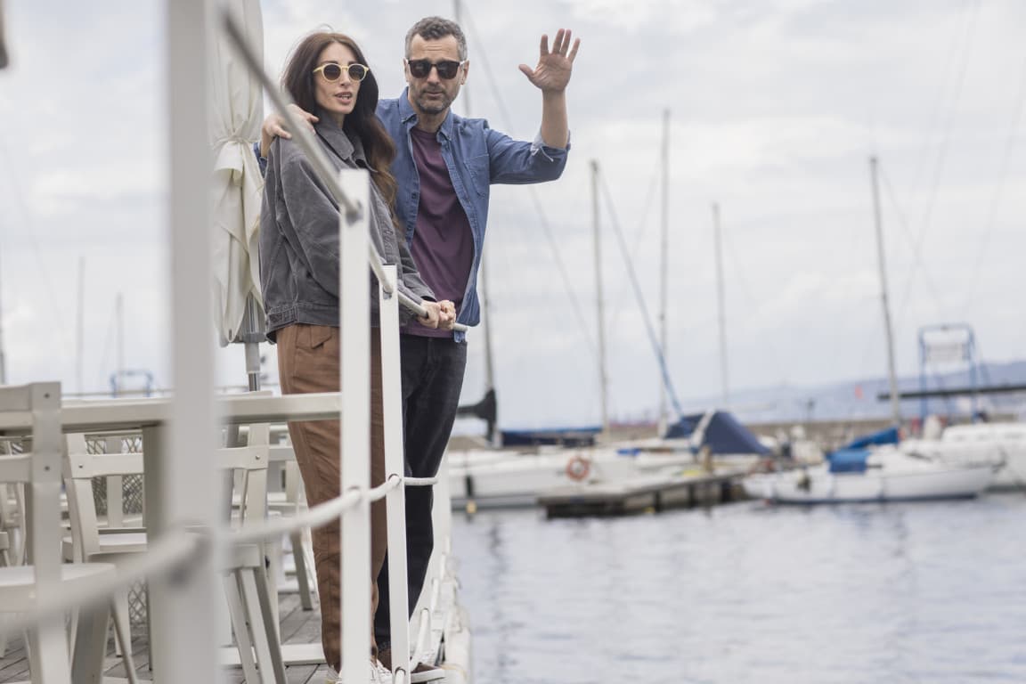 A couple is standing at the harbour in front of the water and the sailboats in Trieste. The guy is waving into the camera while hugging his girlfriend.