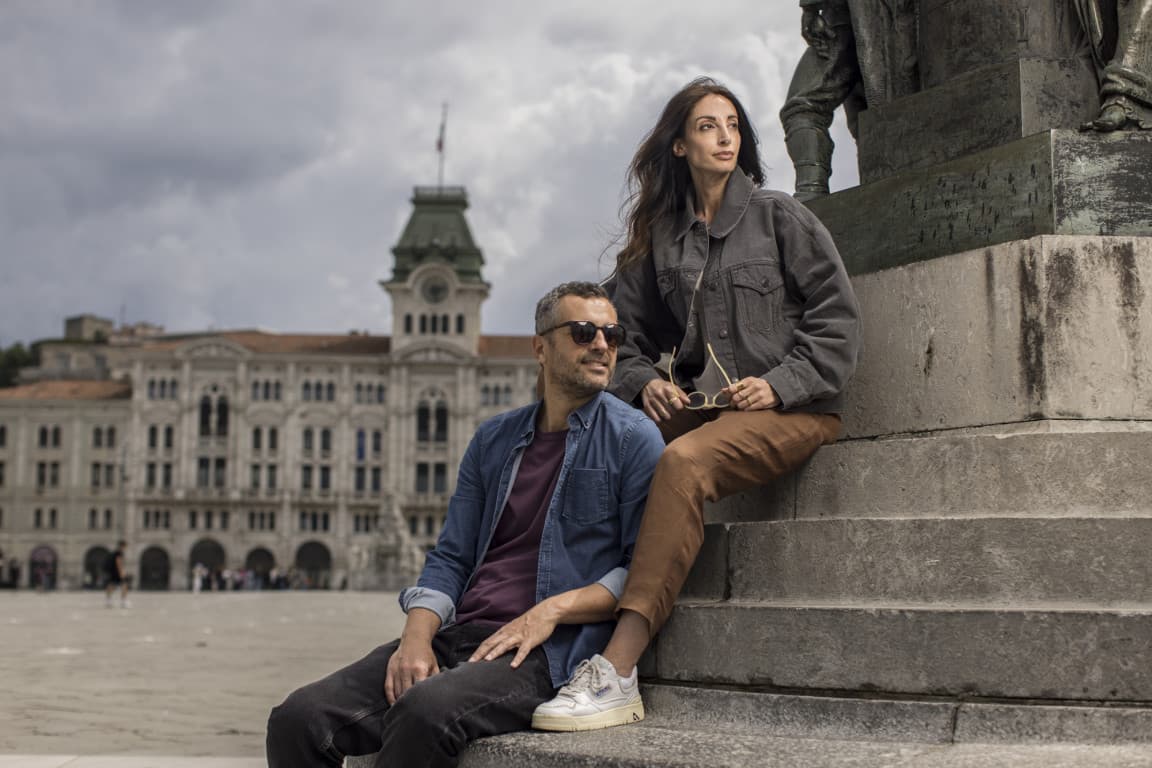 Two tourists are exploring the city of Trieste and take a picture in front of a statue on the main square in the city center.