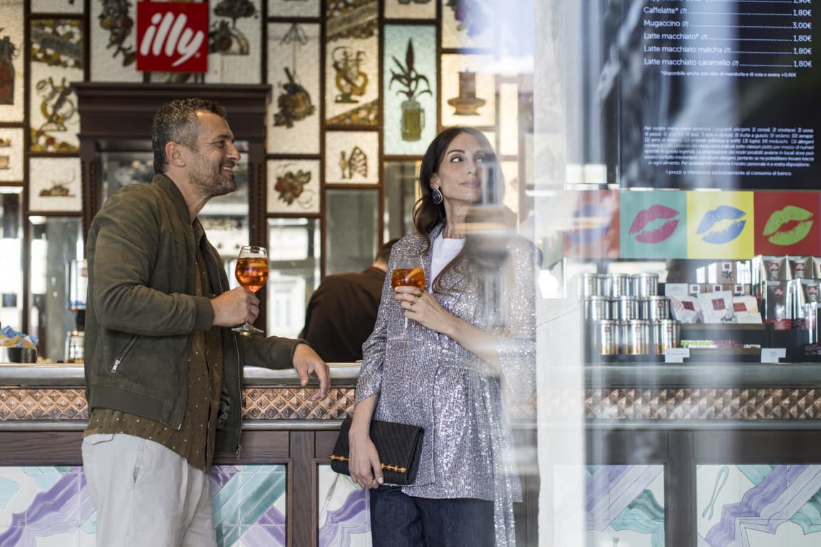 Ein Paar trinkt einen Aperol Spritz in einer Bar in Triest. Sie lehnen an der Bar und blicken auf den Hafen hinaus.