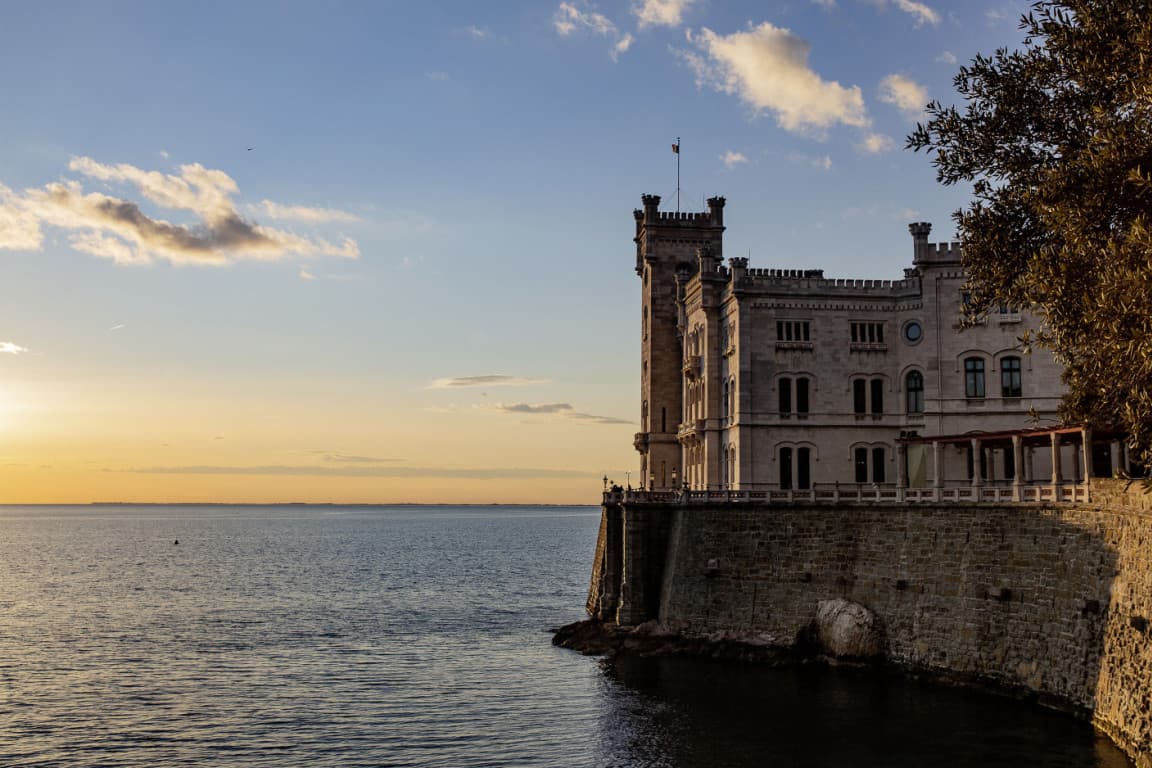 Das Bild zeigt ein markantes Haus am Rande der Klippen an der Küstenlinie von Triest. Es ist in das Sonnenlicht des Sonnenuntergangs getaucht.