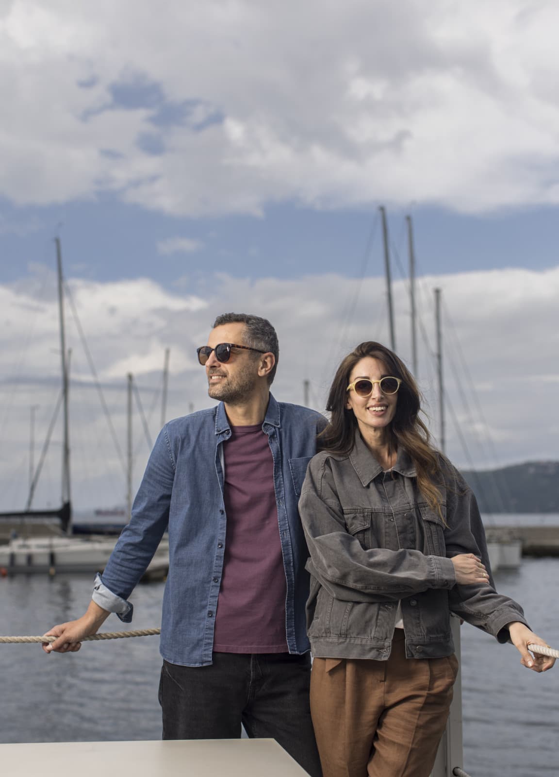 View of a couple posing in front of the harbour in Trieste, leanig against a railing. Both are smiling while the sun is glancing onto their faces.