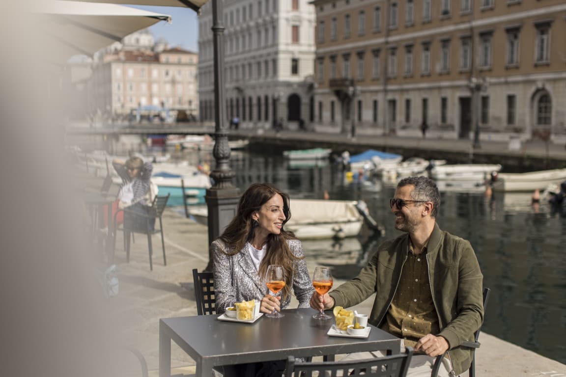 Ein Paar sitzt am Hafen an einem Tisch in der Sonne, genießt ein Getränk und unterhält sich miteinander.
