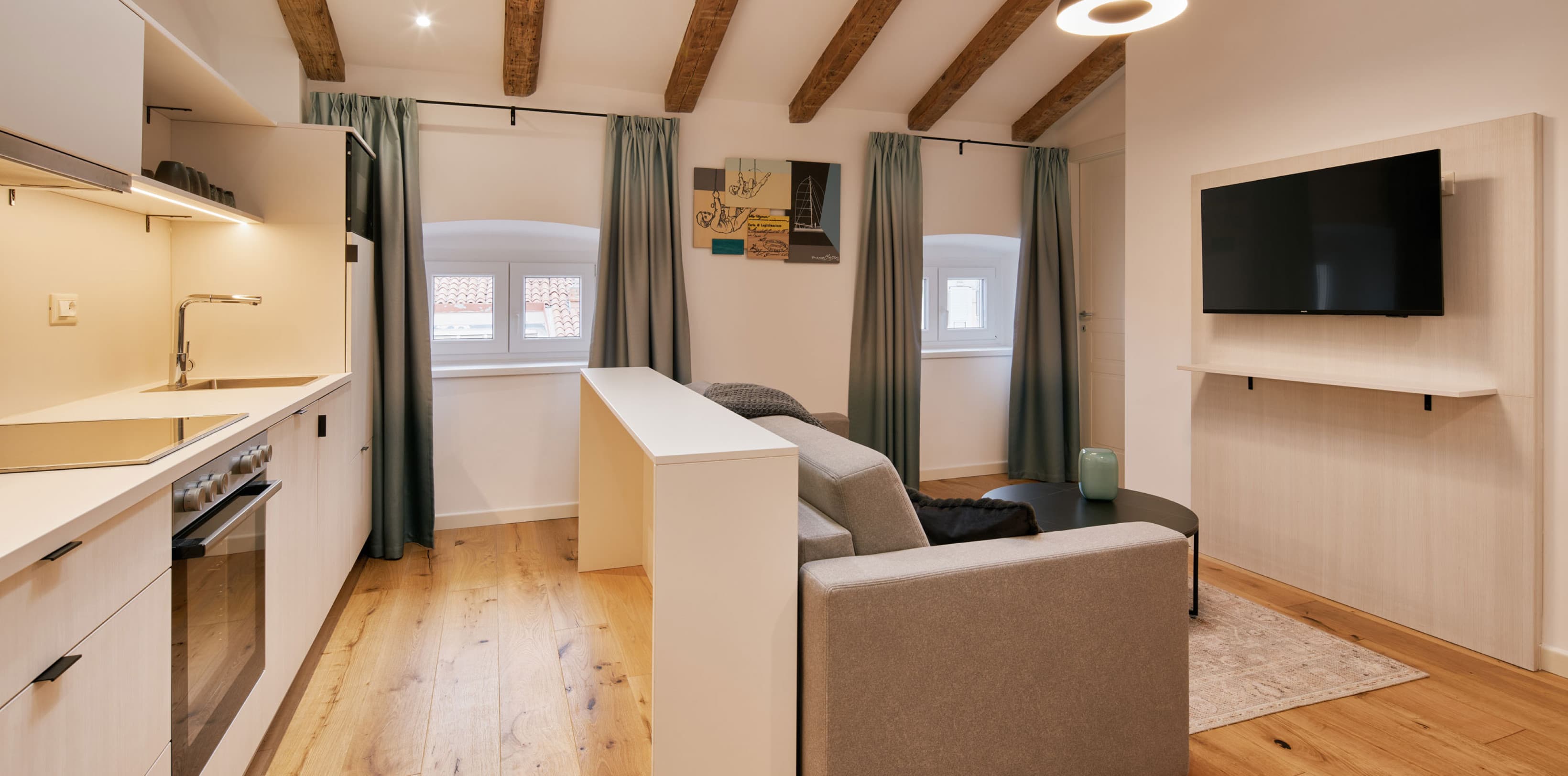 Zu sehen ist das Wohnzimmer des Comfy Studios, mit einer Couch zum Entspannen und einem Fernseher auf der rechten Seite. Auf der linken Seite sehen wir eine voll ausgestattete Küche, die viel Platz bietet, um eine vollständige Mahlzeit wie Frühstück oder Abendessen zuzubereiten.