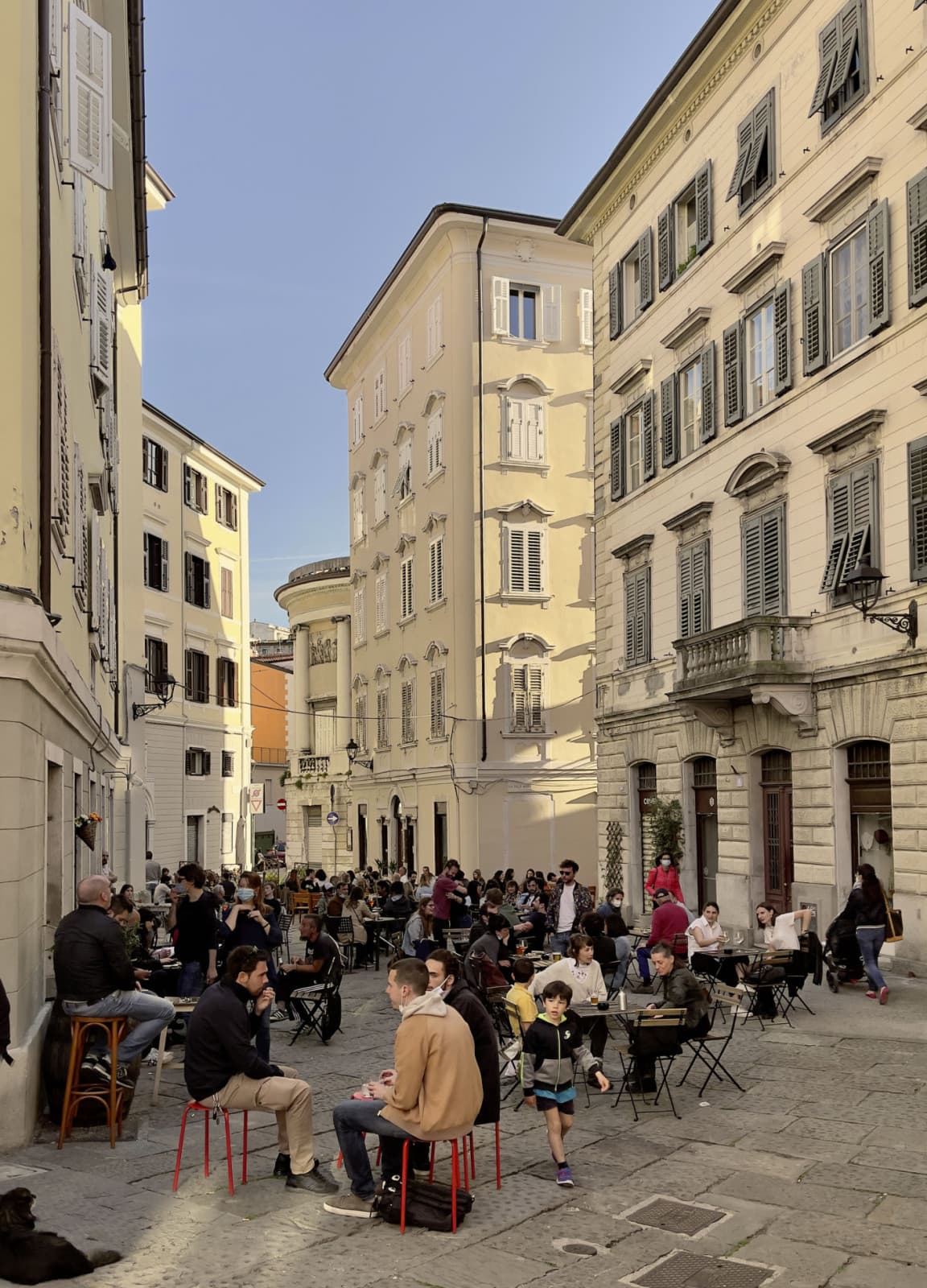 Vista sulla trafficata città di Trieste, dove la gente siede per strada a bere qualcosa tra bellissimi edifici antichi.