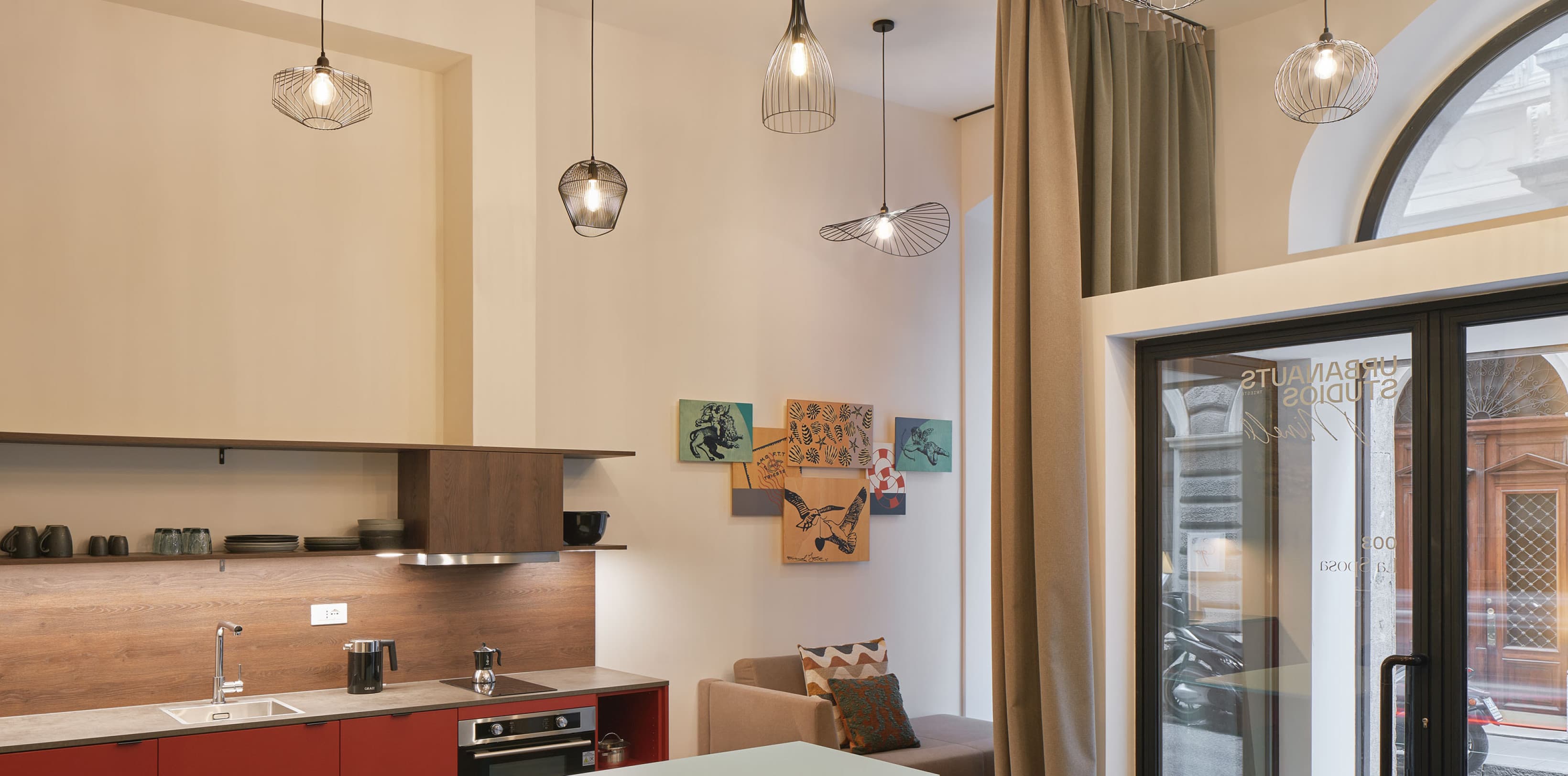 Si vede la zona giorno open space di uno dei loft. La vista è rivolta verso l'ingresso aperto, che si affaccia sulla strada al piano terra. Sullo sfondo si trovano un angolo cottura e un tavolo per cenare, mentre i soffitti alti rendono lo spazio ancora più ampio.