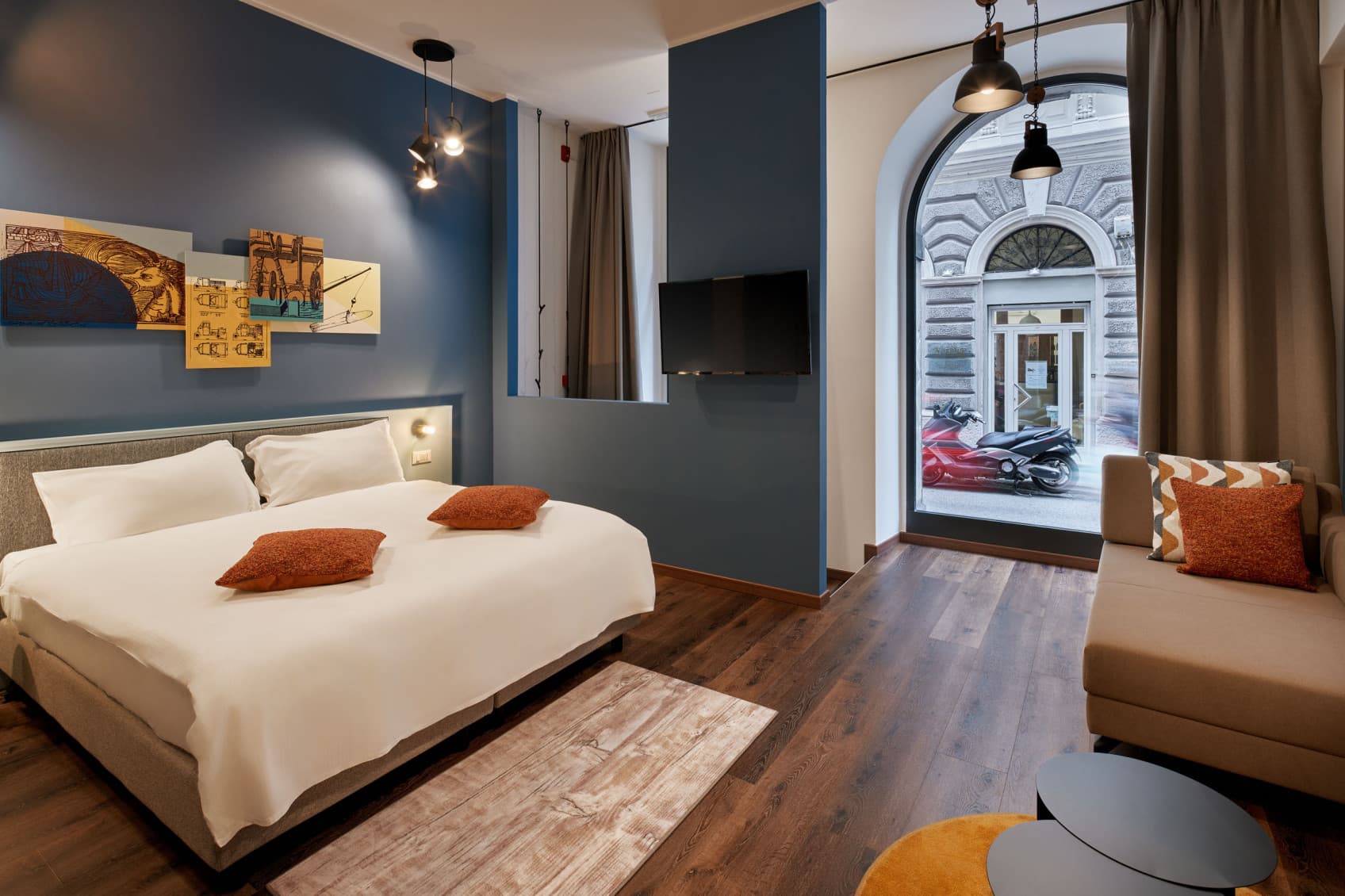 Zu sehen sind das Schlafzimmer und der Wohnbereich, die zusammen einen großen Loft im Erdgeschoss bilden. Auf der linken Seite befindet sich ein großes Doppelbett und auf der rechten Seite des Lofts steht die Couch. Wenn man geradeaus schaut, sieht man das große Schaufenster zur Straße hin.