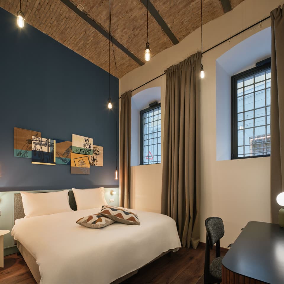 Si vede la camera da letto de Il Farinaio, con un grande letto matrimoniale e una vista sull'appartamento al piano terra. Sullo sfondo si vedono due finestre ristrutturate e lampade appese al soffitto con lunghi fili.